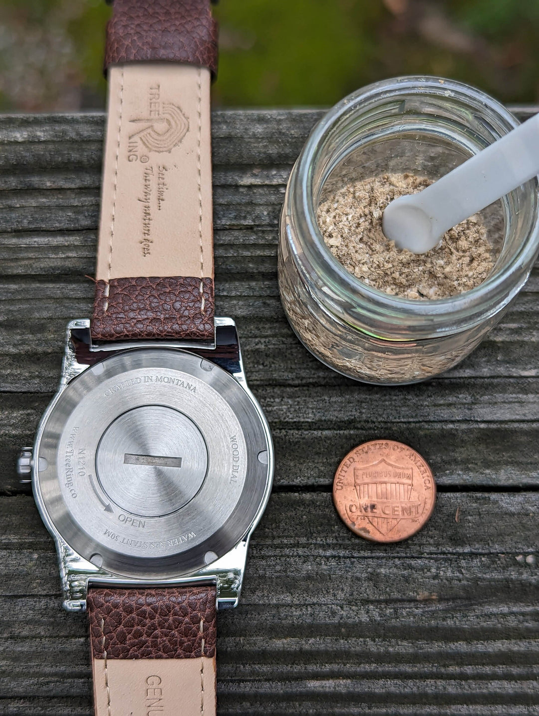 Back view of a Bethlehem olivewood watch, a jar of cremation ashes, and a penny on a wooden surface.